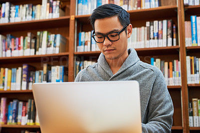 Buy stock photo Shot of a focused young man busy working on his laptop and studying while being seated inside of a library