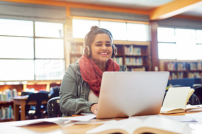 Buy stock photo Portrait of a cheerful young female student working on a laptop while listening to music with her headphones inside of a library