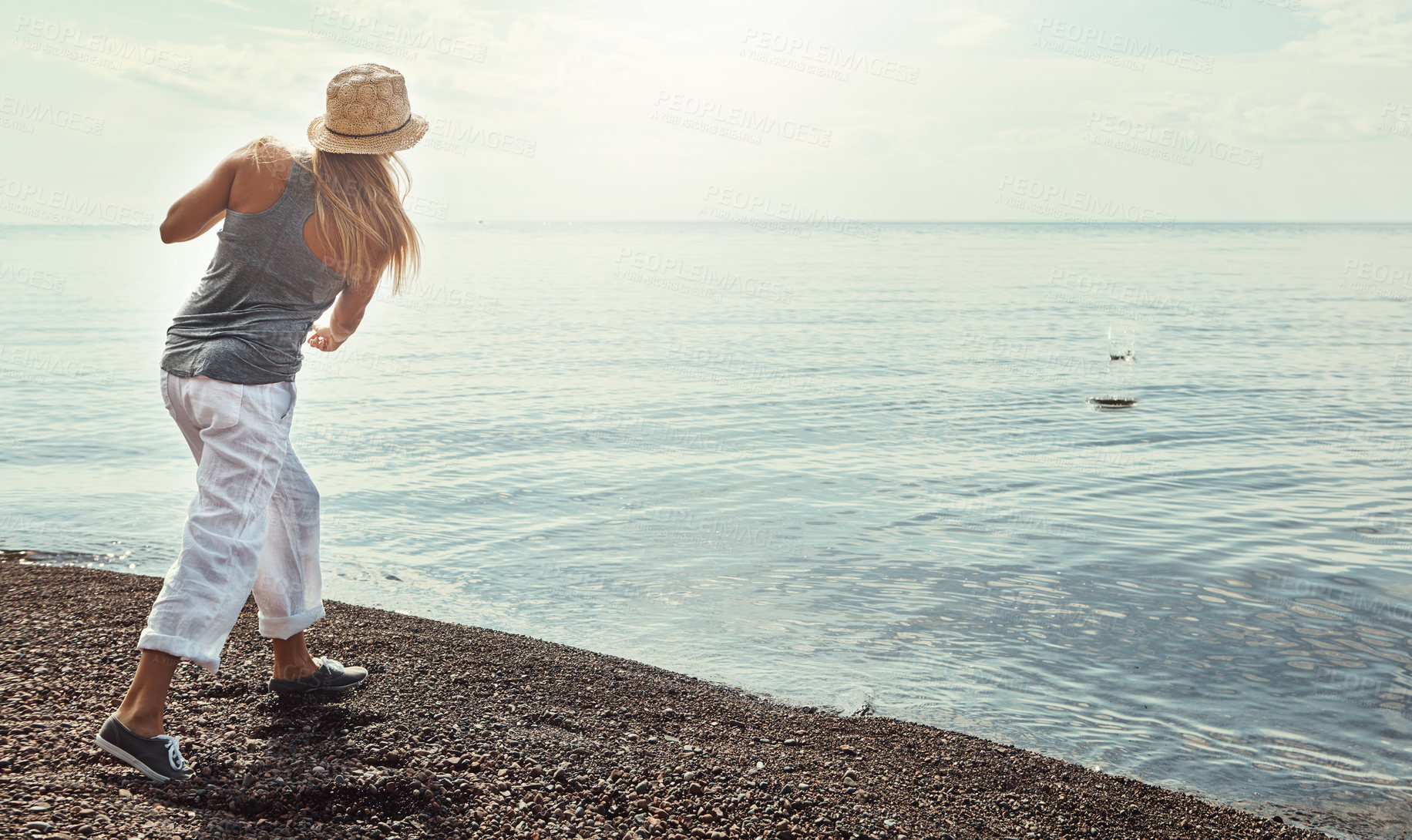 Buy stock photo Beach, ocean and woman skipping rocks outdoor in summer for hobby, leisure or vacation from back. Coast, earth and sky with person throwing stone into sea or water for break, holiday or weekend trip