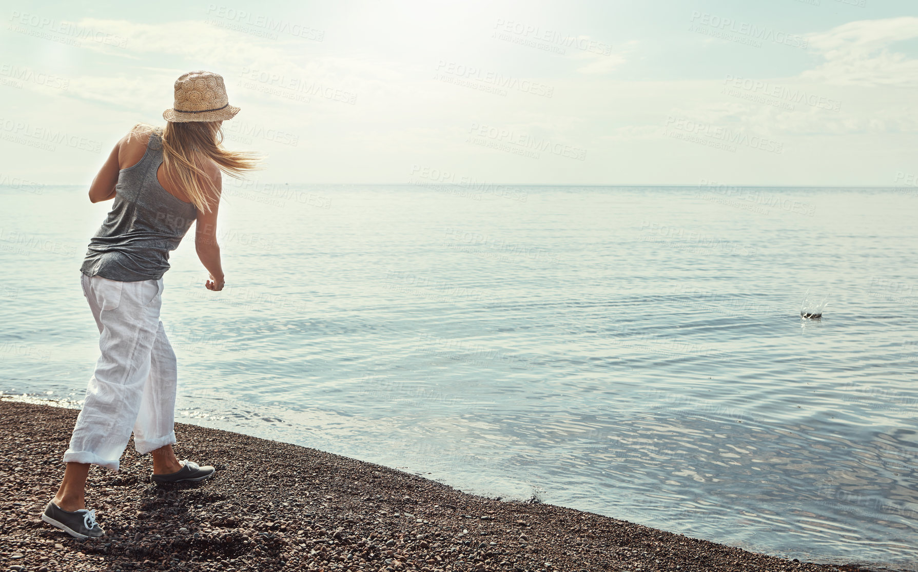 Buy stock photo Beach, water and woman skipping rocks outdoor in summer for hobby, leisure or vacation from back. Coast, ocean and sky with person throwing stone into sea or nature for break, holiday or weekend trip