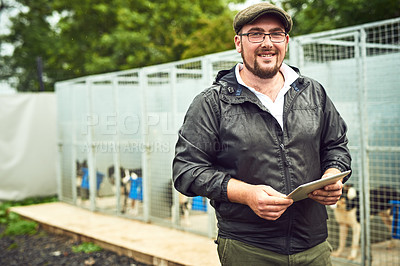 Buy stock photo Portrait of a cheerful young farmer standing with a digital tablet while a group of dogs can be seen relaxing in cages in the background