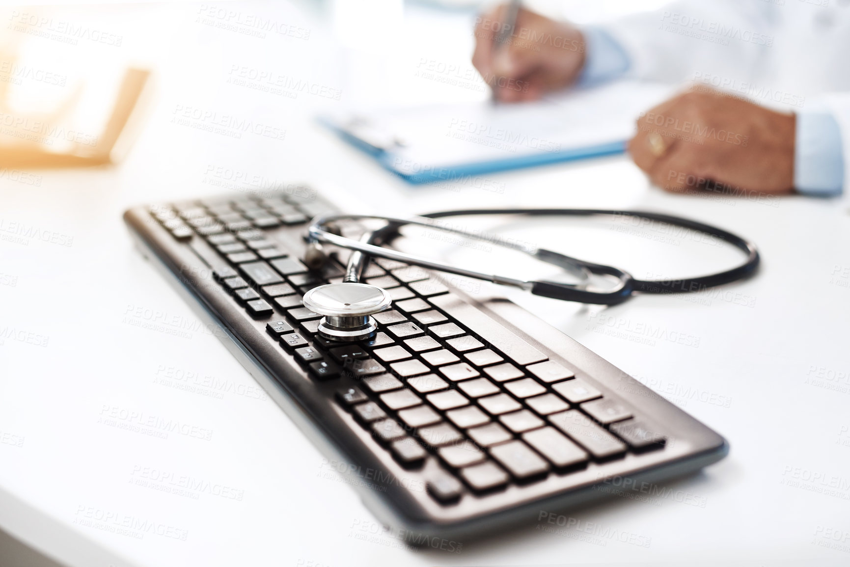 Buy stock photo Shot of a keyboard with a stethoscope on it while an unrecognizable man writes on a form in the background