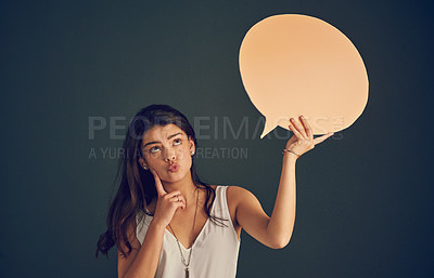 Buy stock photo Studio shot of a carefree young woman holding up a speech bubble while contemplating and standing against a dark background