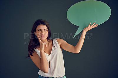 Buy stock photo Studio shot of a carefree young woman holding up a speech bubble while contemplating and standing against a dark background