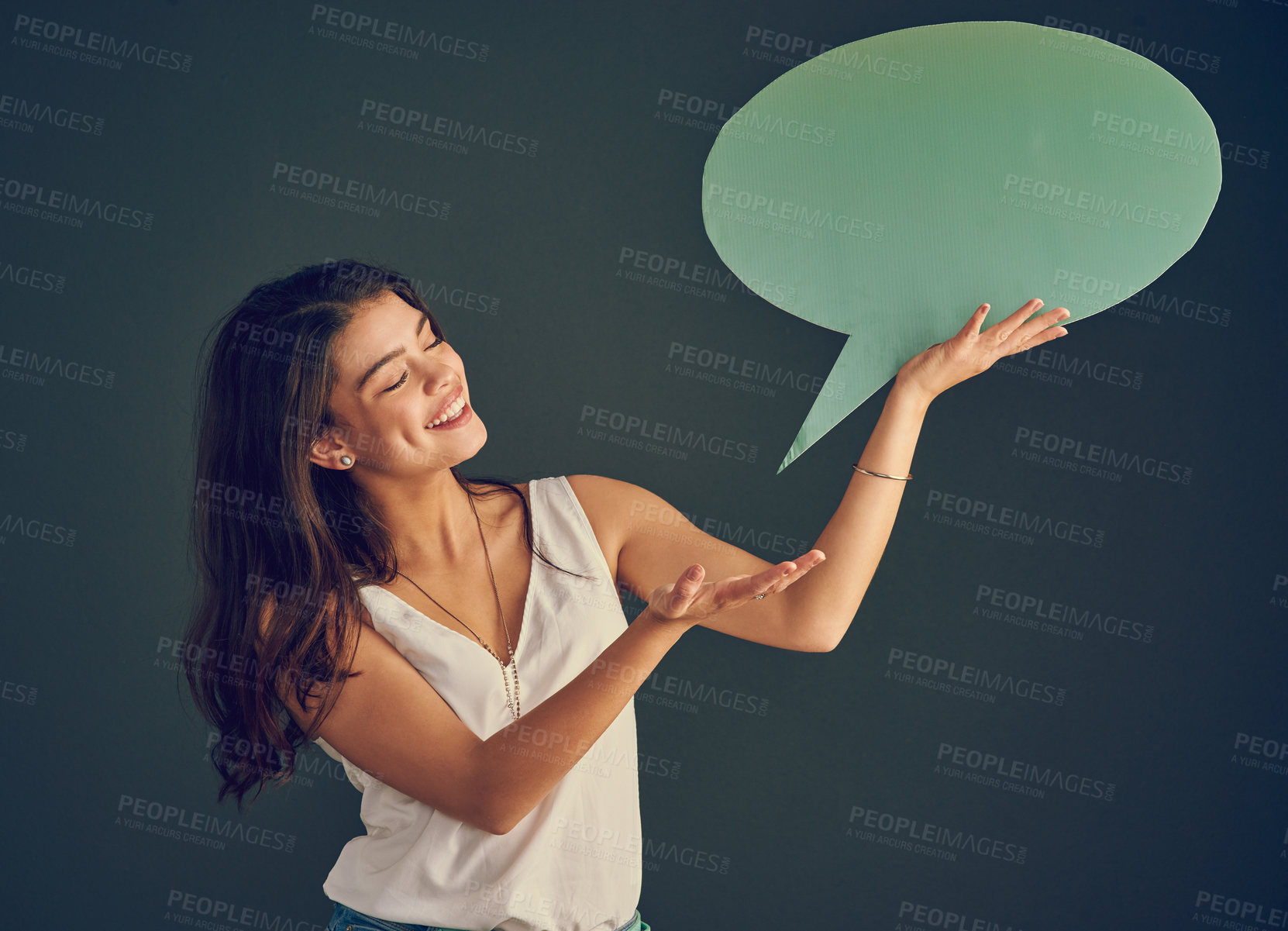 Buy stock photo Studio shot of a cheerful young woman holding up a speech bubble while standing against a dark background