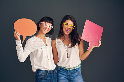 Buy stock photo Studio shot of two cheerful young women holding up speech bubbles while standing against a dark background