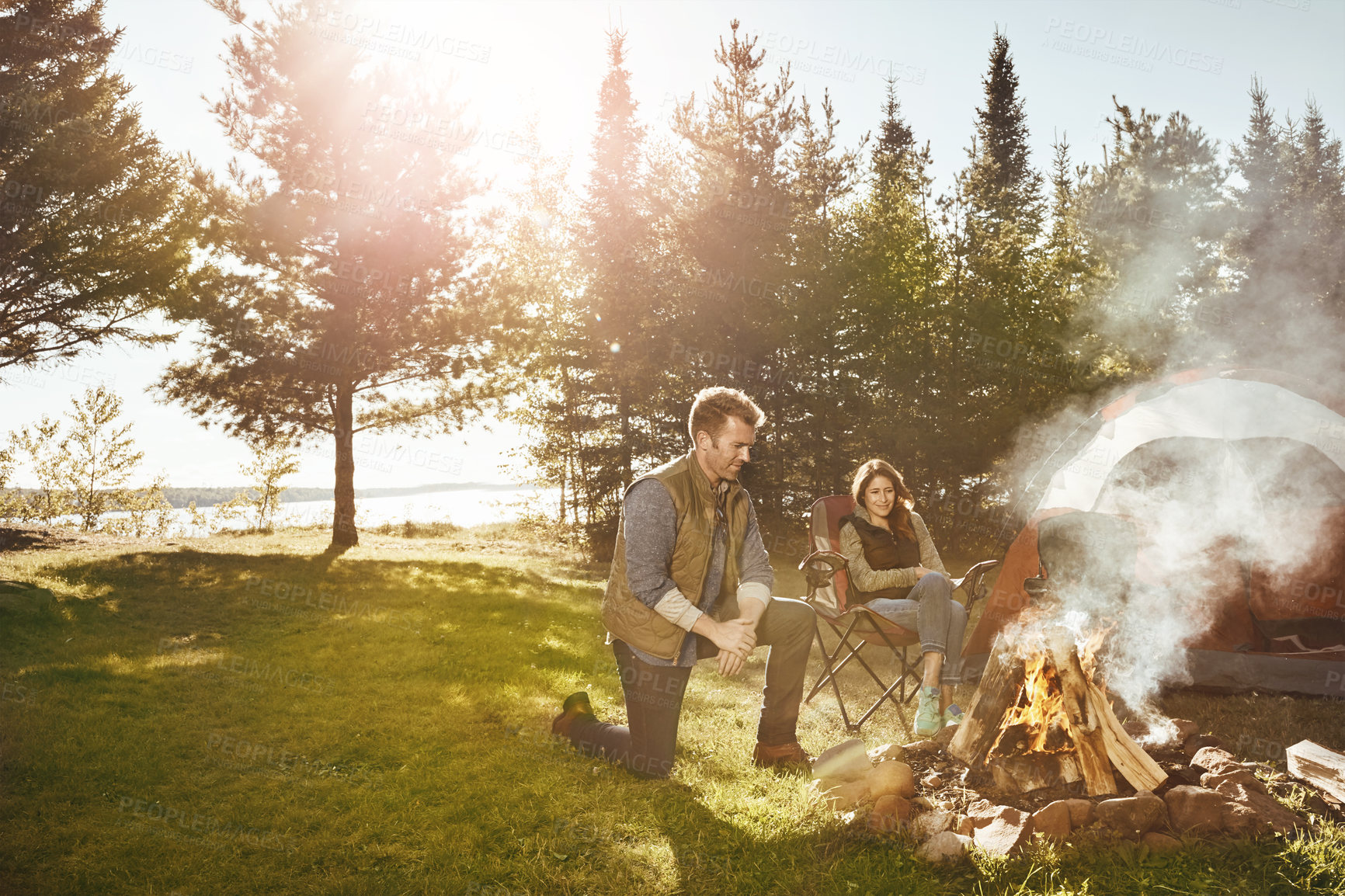 Buy stock photo Nature, fire and couple camping in outdoor forest for vacation, holiday or adventure together. Travel, bonding and young man and woman by flame and tent in woods for weekend trip in New Zealand.