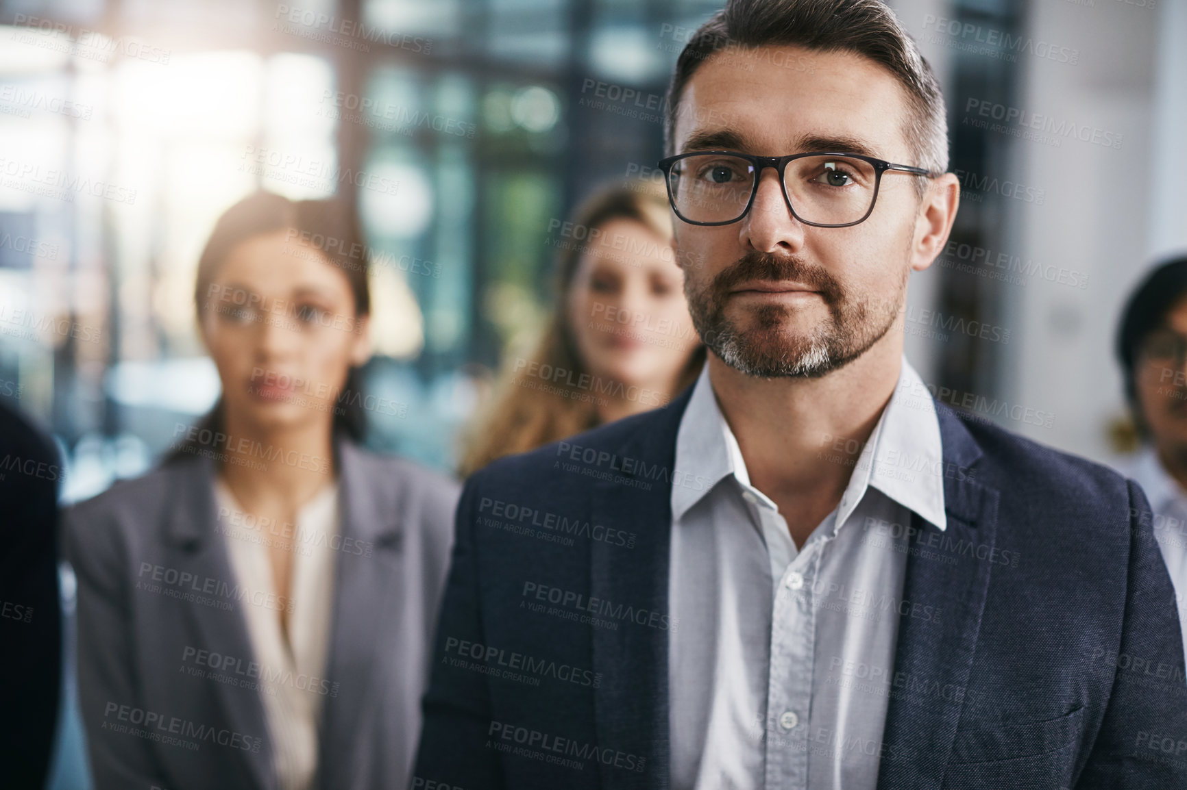 Buy stock photo Portrait of a mature businessman standing in an office with his colleagues in the background