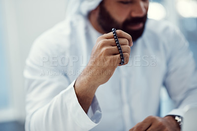 Buy stock photo Cropped shot of a young businessman dressed in Islamic traditional clothing holding prayer beads while working in his office