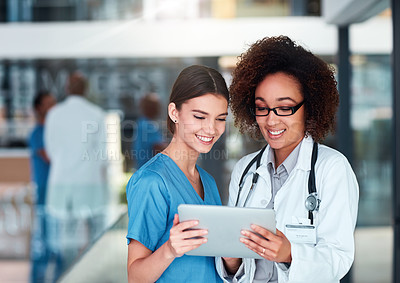 Buy stock photo Shot of two cheerful young doctors working on a digital tablet together while standing in the hospital during the day