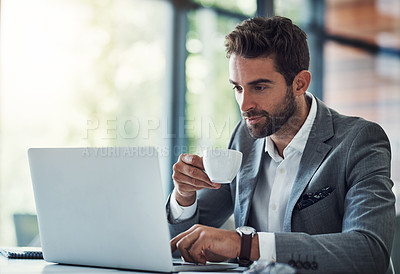 Buy stock photo Shot of a handsome young businessman using a laptop and having coffee at his desk in a modern office