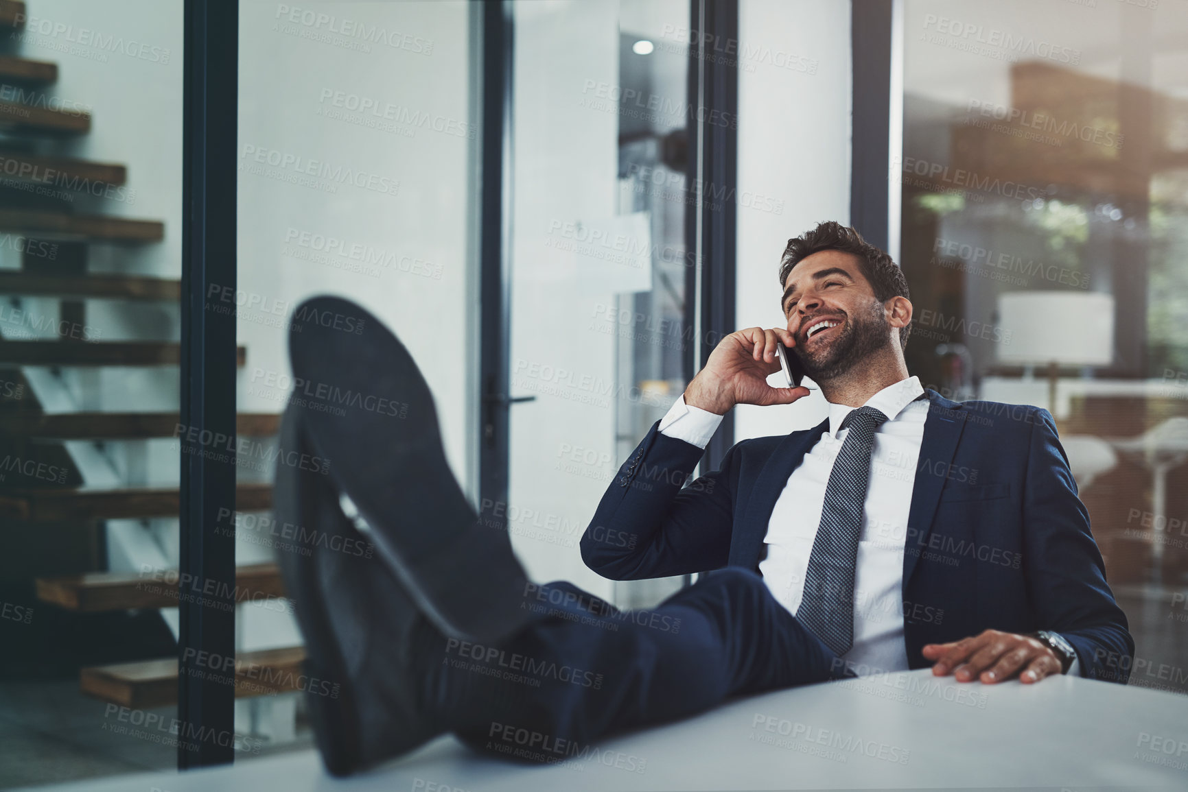 Buy stock photo Shot of a handsome young businessman relaxing with his feet up on an office desk while using his phone