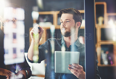 Buy stock photo Shot of a handsome young businessman using a digital tablet while brainstorming on a glass wall in an office