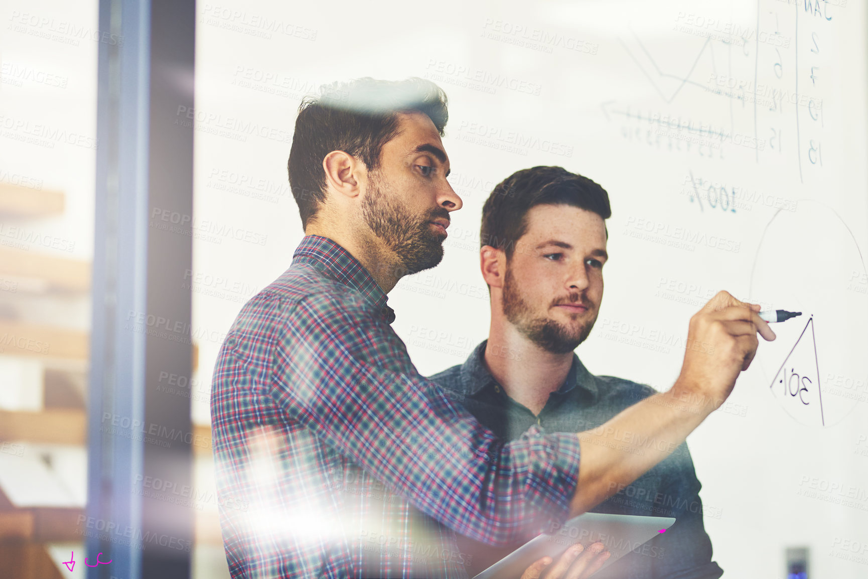 Buy stock photo Shot of two young businessmen using a digital tablet while brainstorming on a glass wall in an office