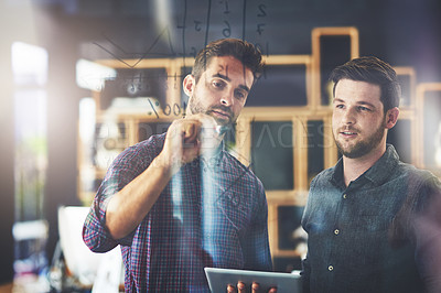 Buy stock photo Shot of two young businessmen using a digital tablet while brainstorming on a glass wall in an office