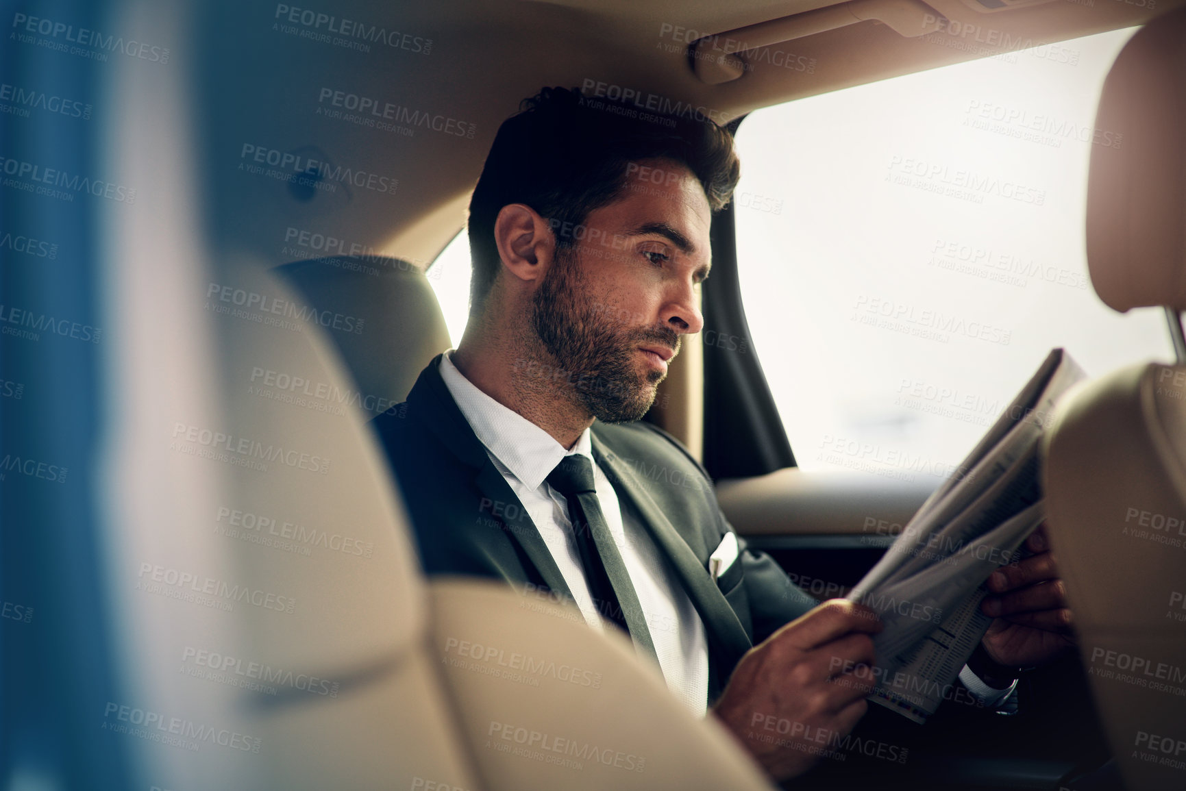 Buy stock photo Shot of a confident young businessman reading the newspaper while being seated in the backseat of a car