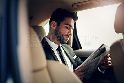 Buy stock photo Shot of a confident young businessman reading the newspaper while being seated in the backseat of a car