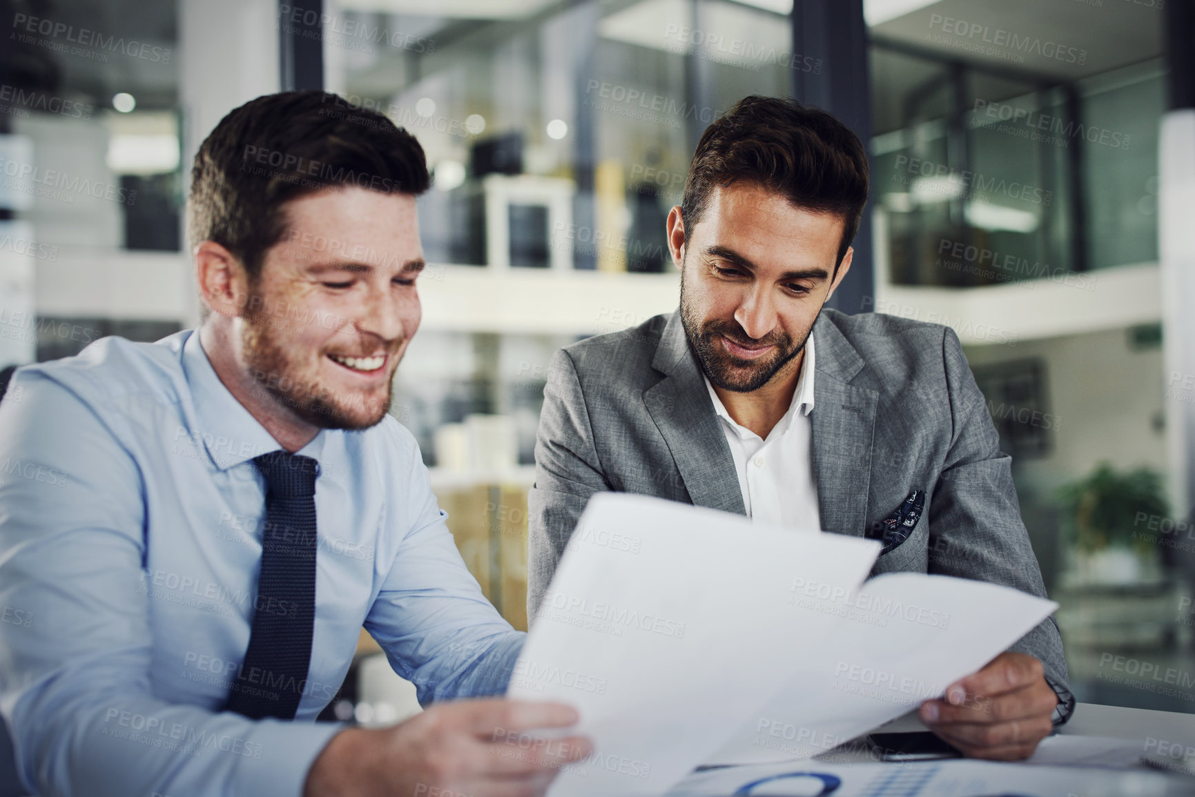 Buy stock photo Shot of two cheerful young businessmen working through business plans together inside of the office