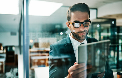 Buy stock photo Shot of a cheerful young businessman browsing on his digital tablet while standing inside of the office