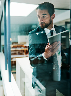 Buy stock photo Shot of a confident young businessman browsing on his digital tablet while standing inside of the office