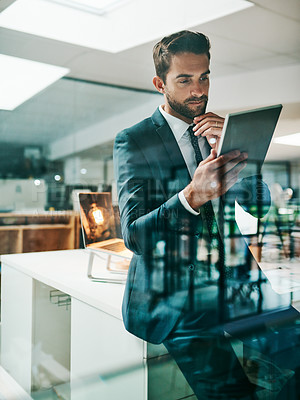 Buy stock photo Shot of a confident young businessman browsing on his digital tablet while standing inside of the office