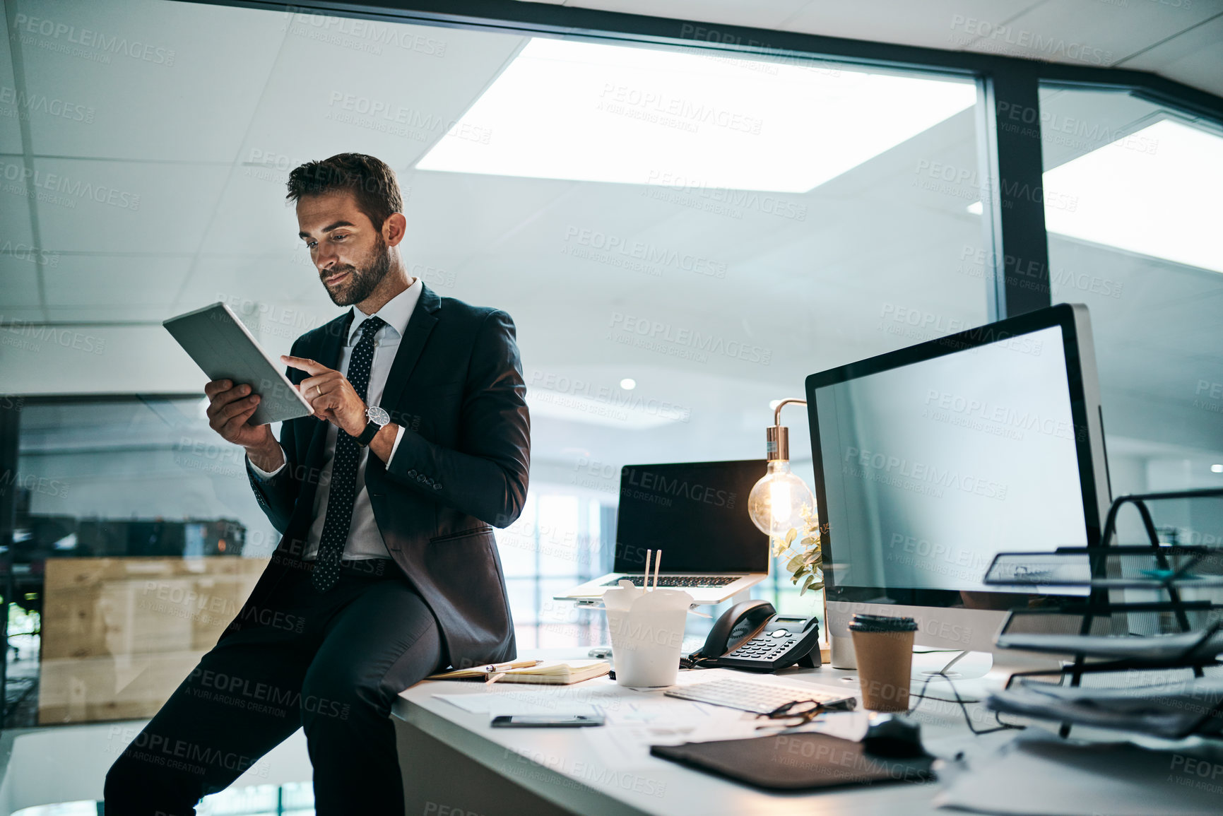 Buy stock photo Shot of a confident young businessman browsing on his digital tablet while standing inside of the office