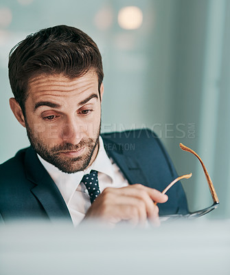 Buy stock photo Shot of a tired young businessman holding a pair of glasses while working on a computer at the office
