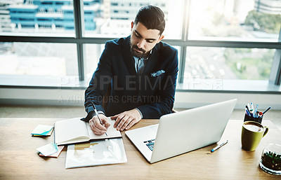 Buy stock photo Shot of a young businessman writing in a notebook at his desk in a modern office