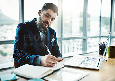 Buy stock photo Shot of a young businessman writing in a notebook at his desk in a modern office