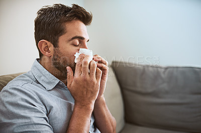 Buy stock photo Shot of a uncomfortable looking young man sneezing into a tissue while being seated on a couch at home