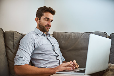 Buy stock photo Shot of a focused young man working on his laptop while being seated on a couch at home during the day