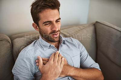 Buy stock photo Shot of an uncomfortable looking young man holding his chest in discomfort while being seated on a couch at home