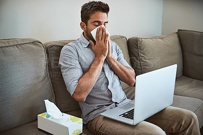 Buy stock photo Shot of a uncomfortable looking young man sneezing into a tissue while trying to work on his laptop at home