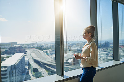 Buy stock photo Shot of a confident young businesswoman drinking coffee while standing next to a window and looking through it inside of the office