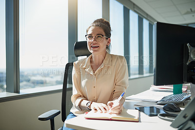 Buy stock photo Shot of a cheerful young businesswoman writing in her notebook while being seated at her desk inside the office during the day