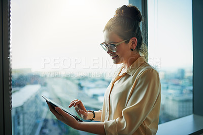 Buy stock photo Shot of a young businesswoman browsing on a digital tablet while standing next to a window in the office