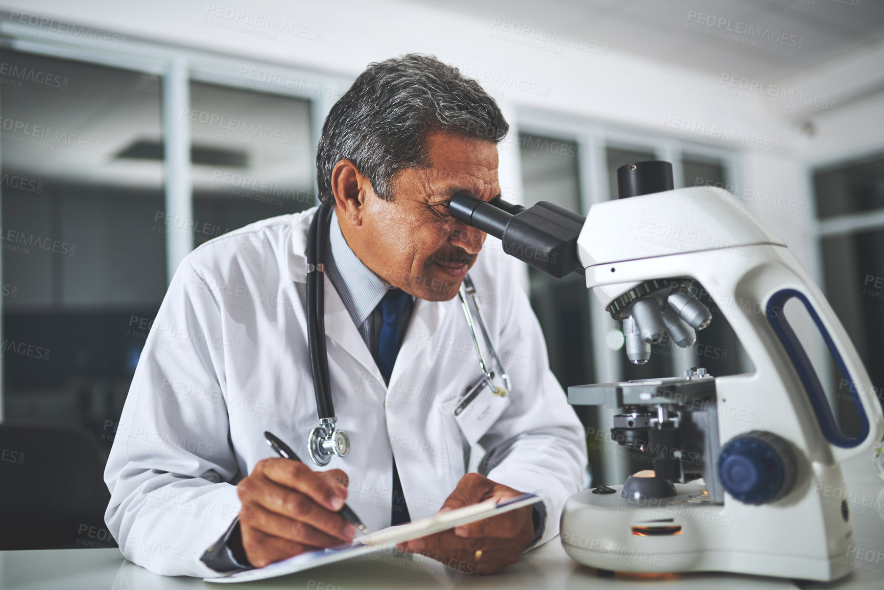 Buy stock photo Shot of a mature scientist using a microscope and recording his findings in a laboratory