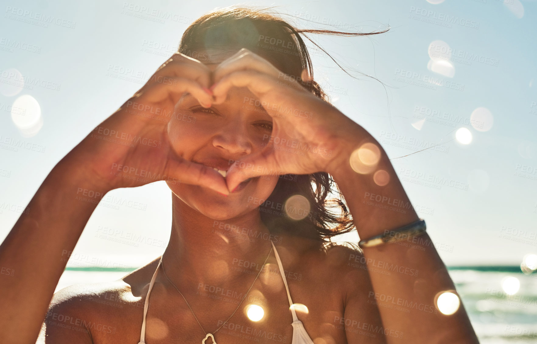 Buy stock photo Portrait of a beautiful young woman making a heart shaped gesture with her hands at the beach