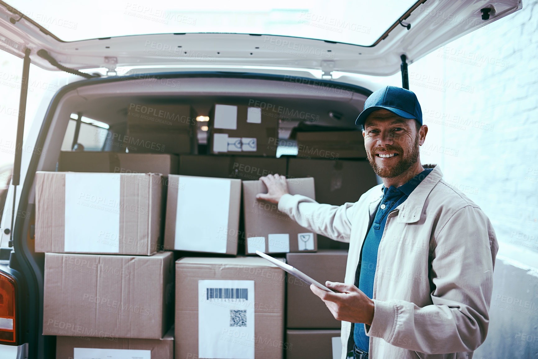 Buy stock photo Portrait of a cheerful young delivery man standing next to a van full of boxes while holding a digital tablet