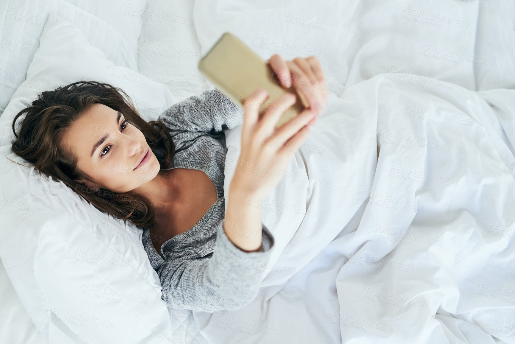 Buy stock photo Shot of an attractive young woman typing on her cellphone while lying in bed at home during the day