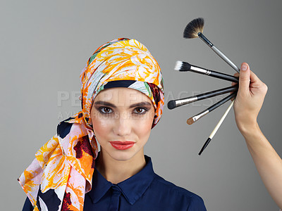 Buy stock photo Studio portrait of a confident young woman wearing a colorful head scarf while looking at different makeup brushes being held in front of her