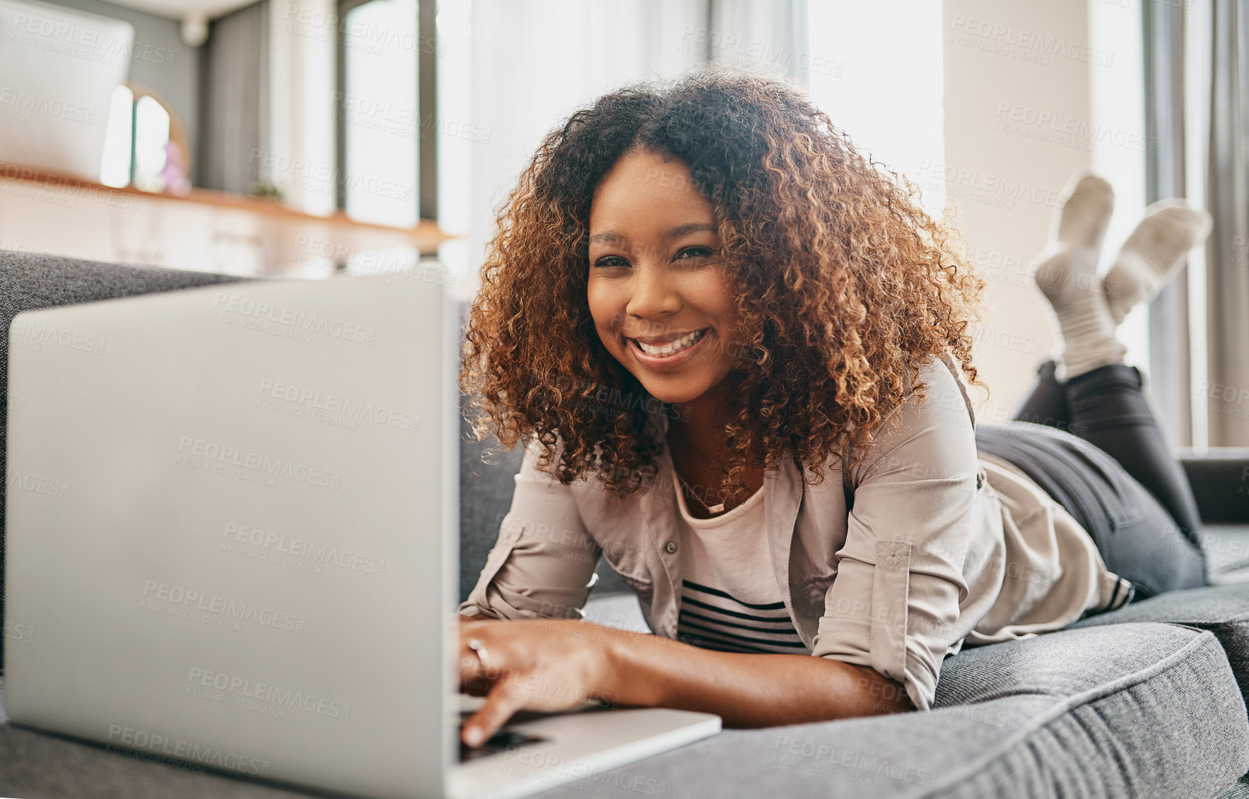 Buy stock photo Shot of a cheerful young woman working on a laptop while lying on a couch at home during the day