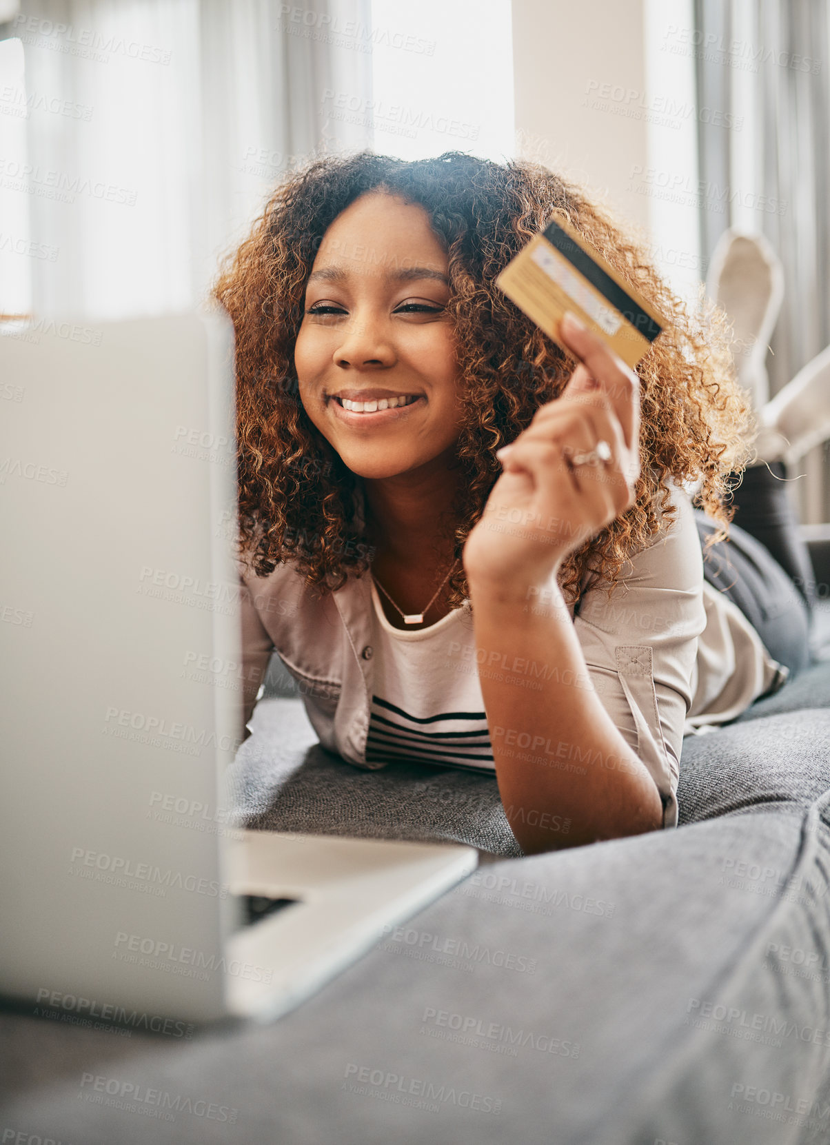 Buy stock photo Shot of a cheerful young woman doing online shopping on her laptop while lying on a couch at home during the day