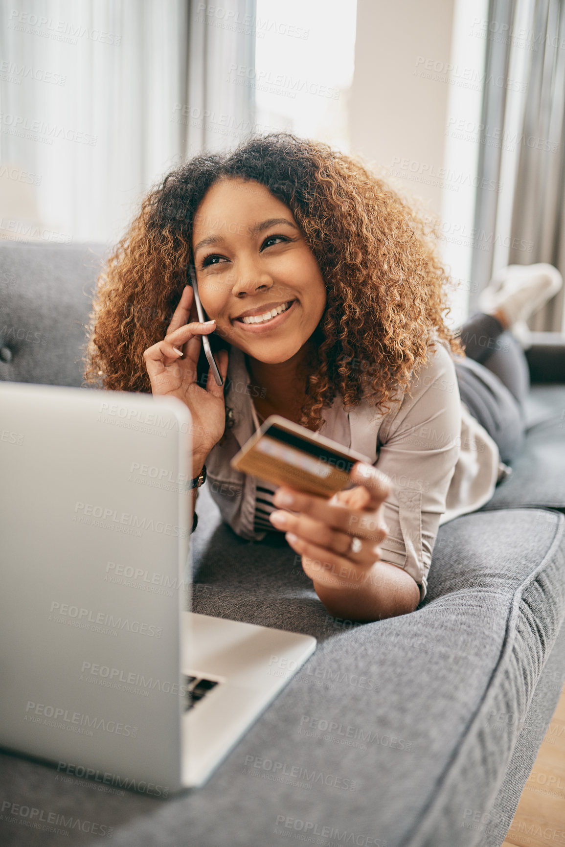 Buy stock photo Shot of a cheerful young woman doing online shopping on her laptop while talking on her cellphone at home