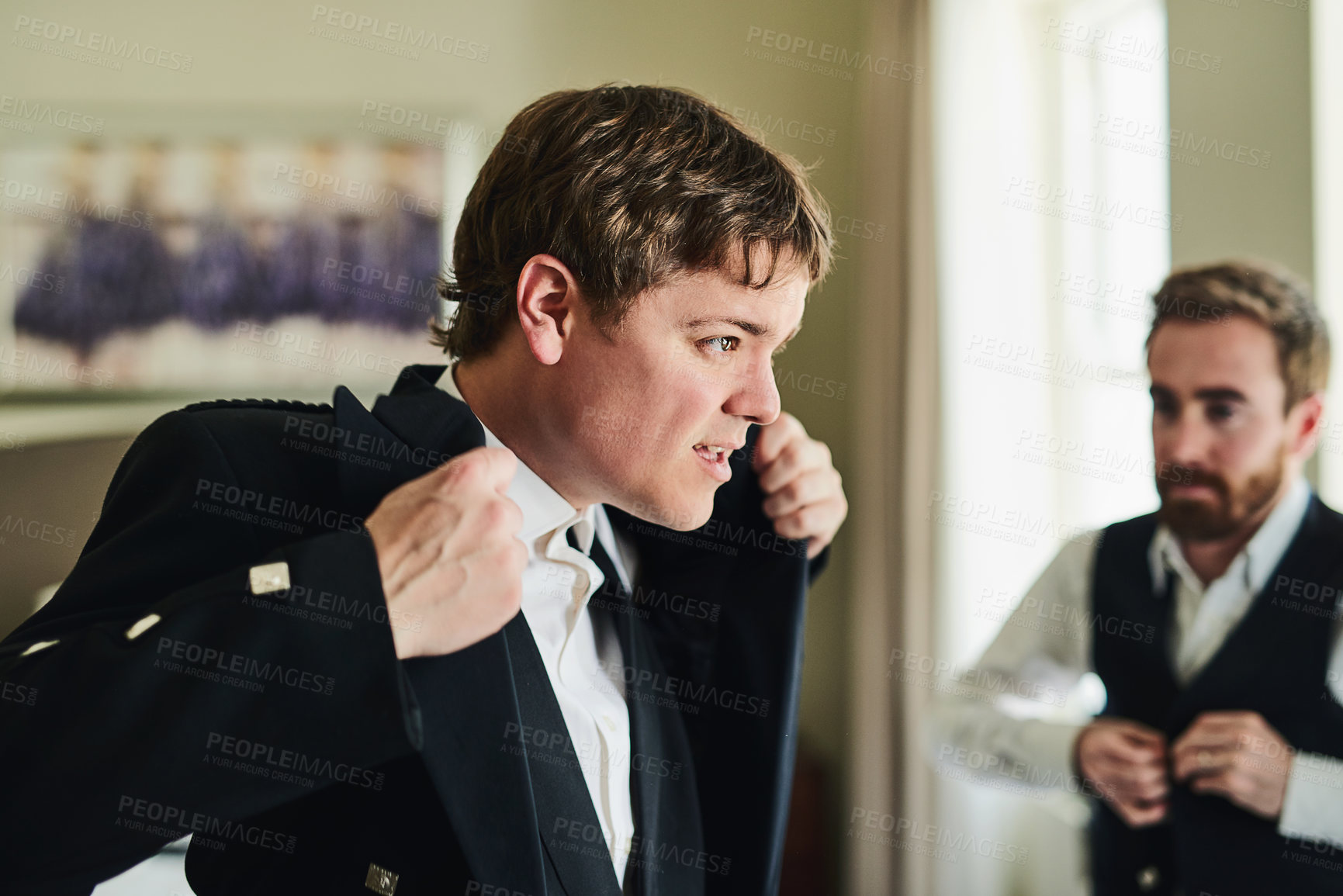 Buy stock photo Shot of a confident young man putting on a suit jacket while looking into a mirror to get ready for a wedding