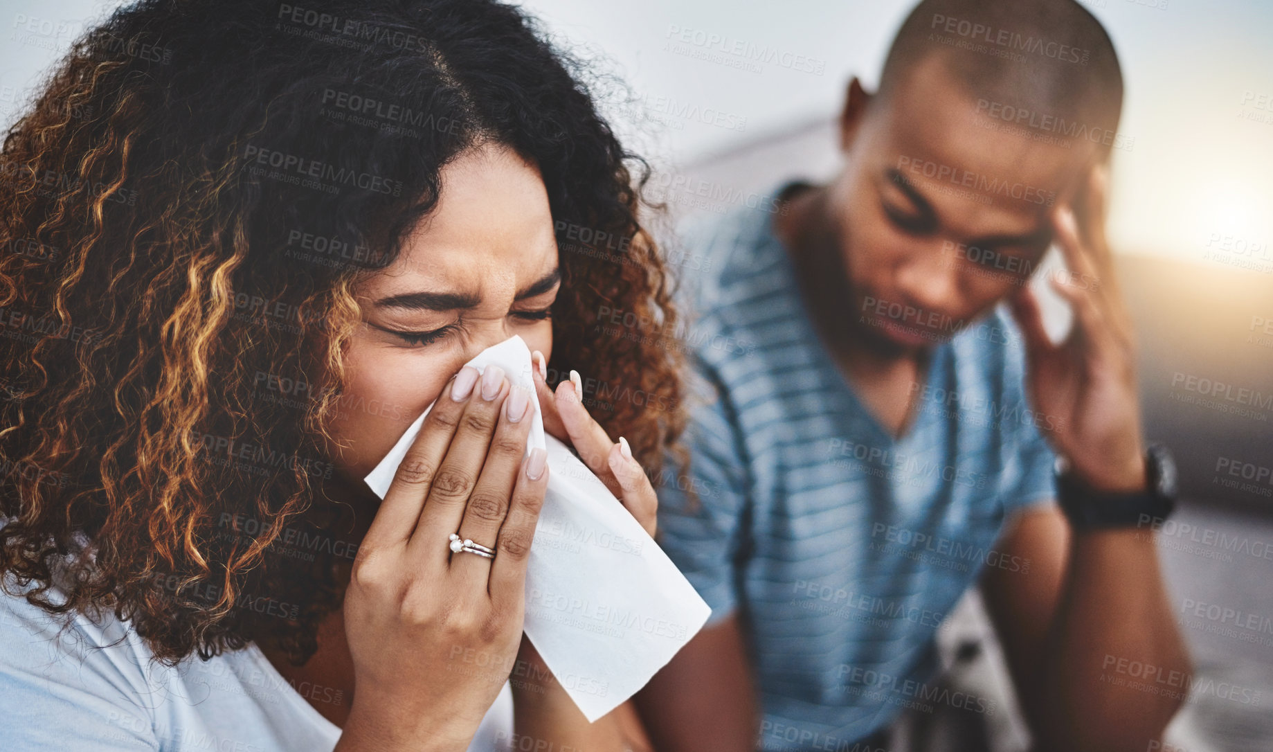 Buy stock photo Shot of a young woman blowing her nose with her boyfriend looking irritated in the background
