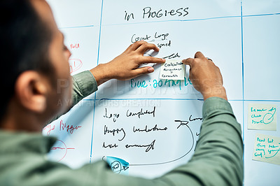 Buy stock photo Over the shoulder shot of a focused young businessman putting a sticky note on a white board in the office during the day