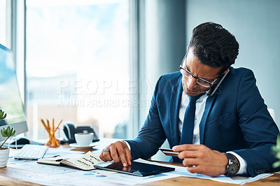 Buy stock photo Shot of a focused young businessman seated at his desk while talking on the phone and browsing on a digital tablet in the office