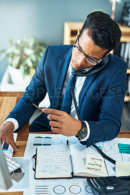 Buy stock photo Shot of a focused young businessman seated at his desk while taking on the phone and typing on a keyboard in the office