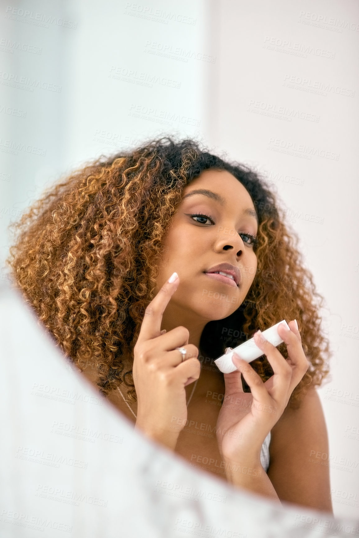 Buy stock photo Shot of a confident young woman applying skin cream on her face while looking into the mirror in the bathroom at home